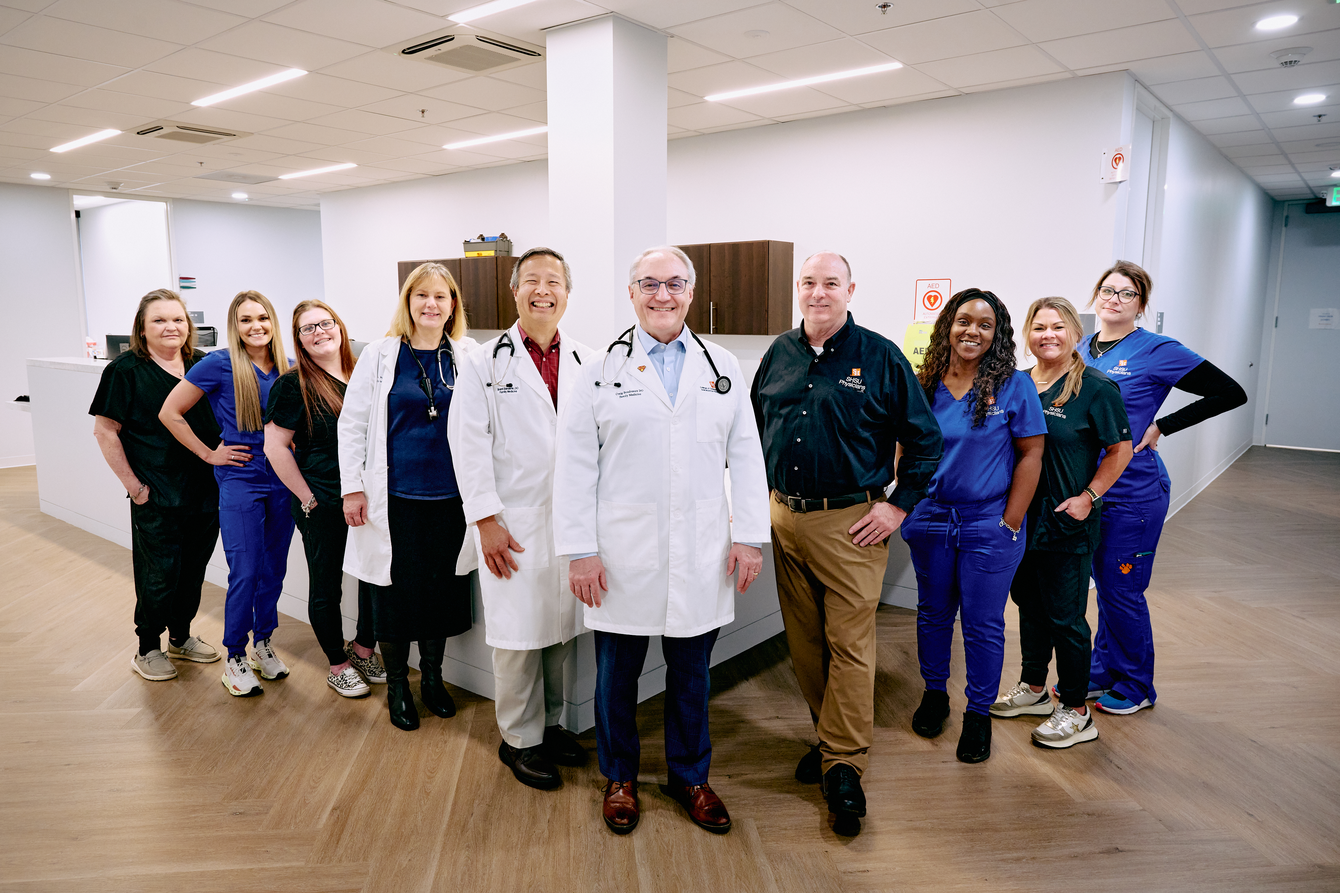 SHSU Physicians doctors and clinic staff stand together in the clinic hallway, smiling as a group in front of the nurses’ station.